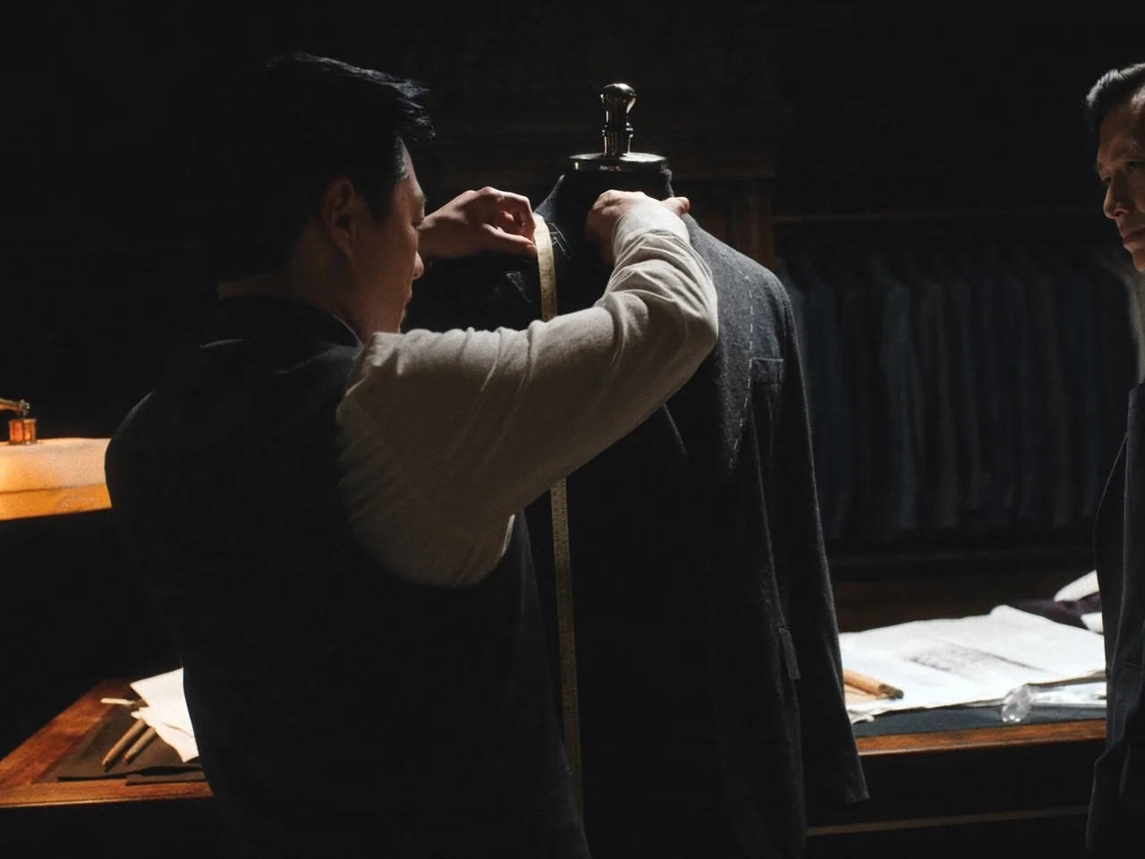 A tailor measuring a bespoke jacket on a mannequin while the client watches, a rack of ready-made suits in the background.